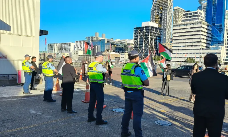 Police officers and several people stand on a city street with tall buildings in the background. Some individuals hold Palestinian flags, and traffic cones create a barrier in the area. It appears to be a public gathering or protest.