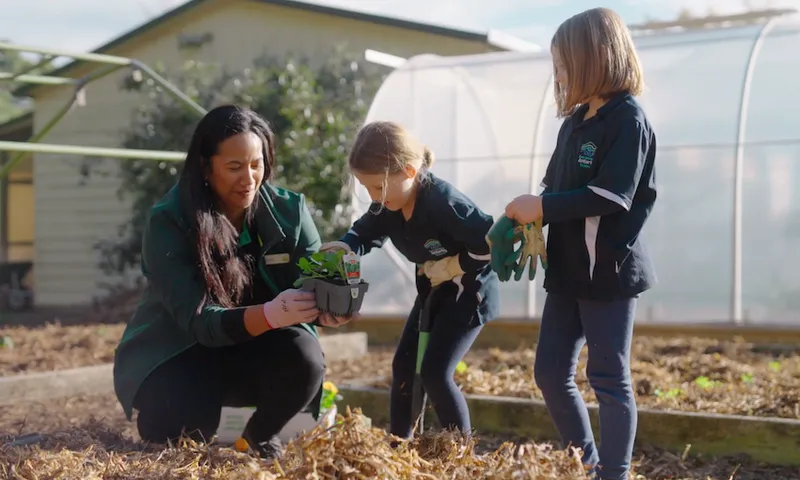 a woman with brown skin and long brown hair holds a seedling while two young girls with light brown hair watch on
