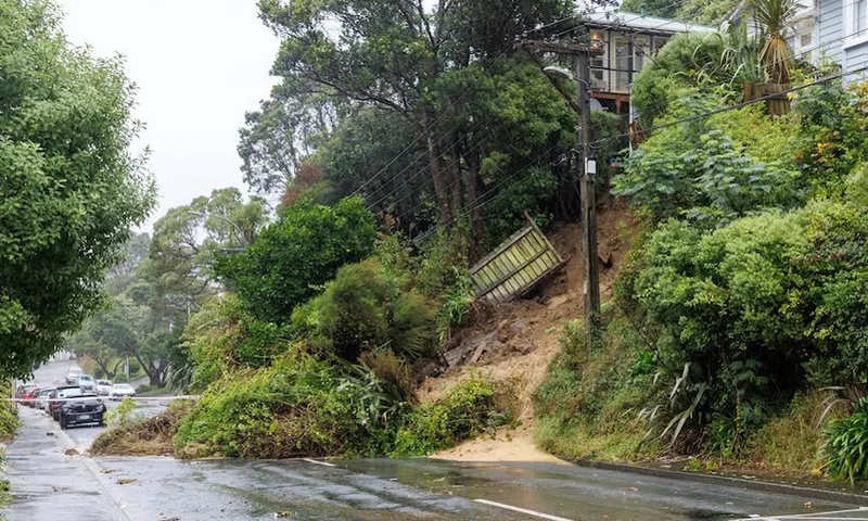 A landslide has caused mud, debris, and parts of a fence to slide onto a wet street, blocking one lane. Trees and vegetation are scattered, with a house partially visible above the slope. Parked cars line the street.