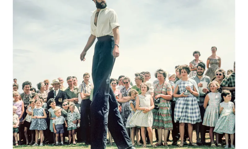 A crowd of people gather to watch a stilt walker. The photo is clearly historic by the clothes which look 1950s.