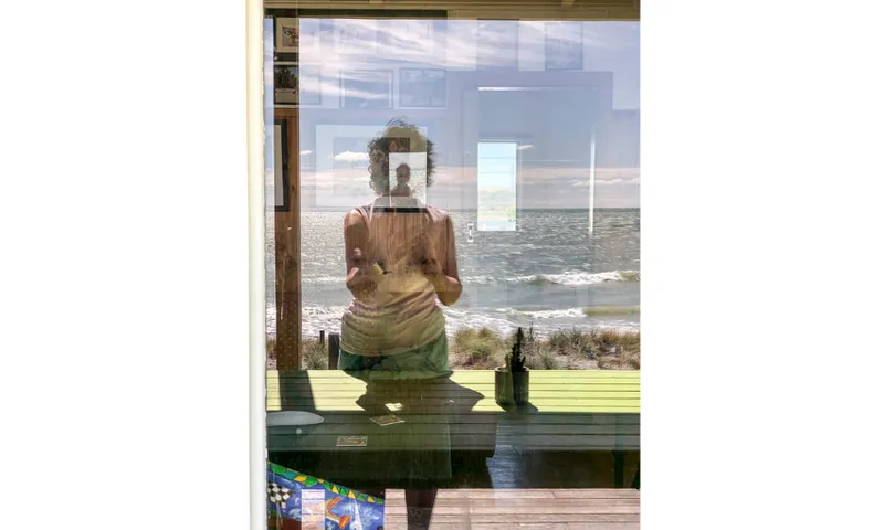 A woman taking a photo is reflected in the window which is looking out towards a beach.