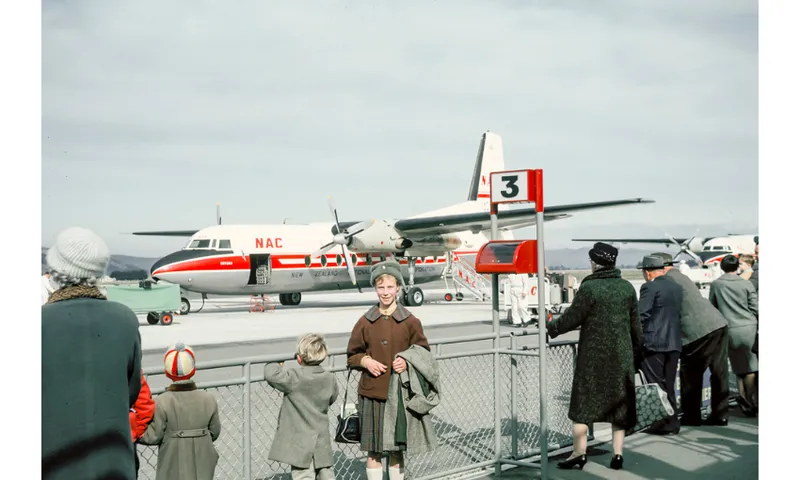 A young girl stands in front of a fench in front of a small aircraft at an airport.