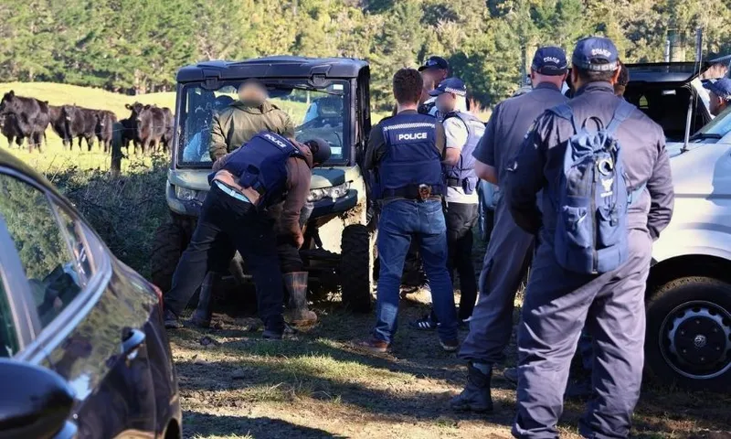 A group of police officers stand around a person working on an off-road vehicle in a rural area, with several cows and trees visible in the background.