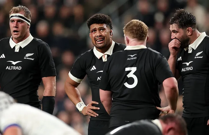 four men in black rugby uniforms stand, puffed, with a large crowd behind them