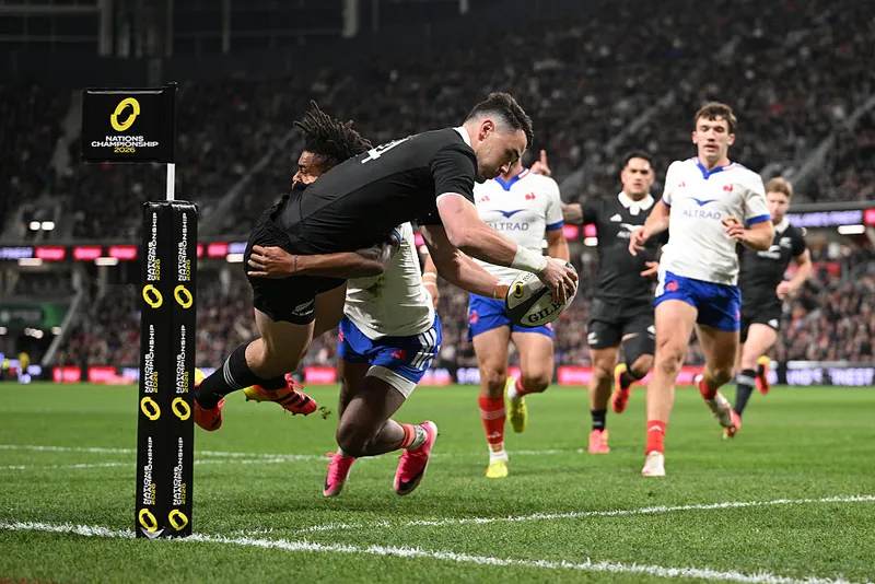 rugby players on a large field at night watch on as a man in a black uniform dives high into the air to score a try while a player in white and blue tackles him