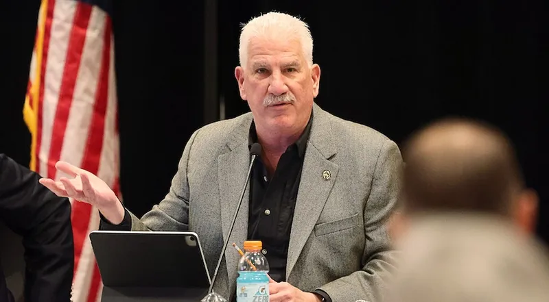 A man with white hair and a mustache sits at a table, speaking and gesturing with one hand. He wears a gray blazer over a black shirt. An American flag and a tablet are visible, along with a bottle in the foreground.