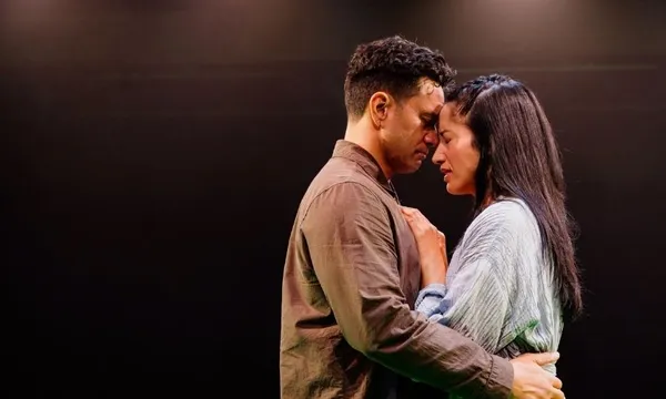 A man and a woman stand close together on a dimly lit stage, gently touching foreheads and holding each other tenderly, conveying a moment of deep emotion and connection.