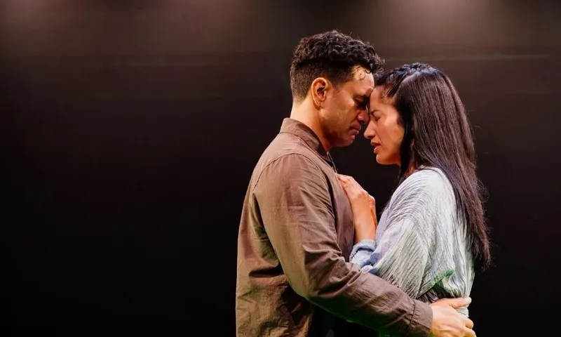A man and a woman stand close together on a dimly lit stage, gently touching foreheads and holding each other tenderly, conveying a moment of deep emotion and connection.