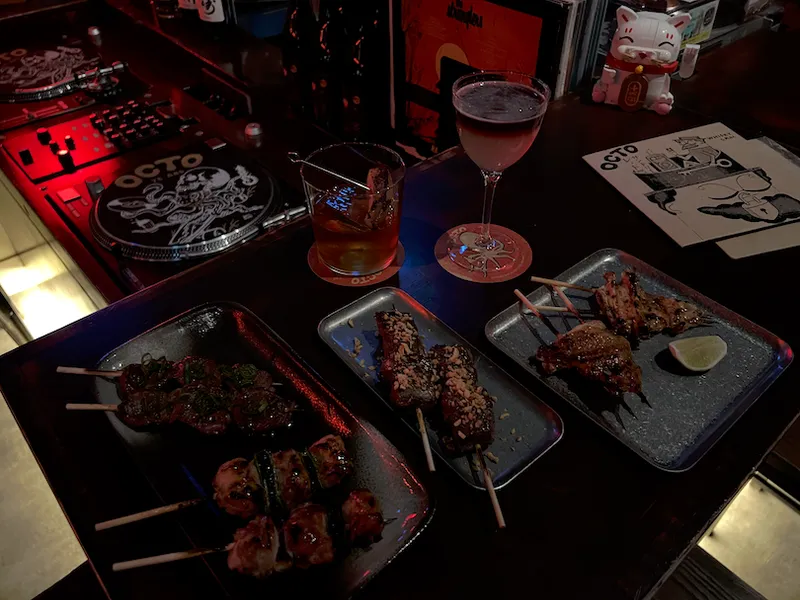 A dimly lit bar counter with vinyl records, two cocktails, and three plates of assorted grilled skewers. A menu, sake bottles, and a beckoning cat statue are also visible in the background.