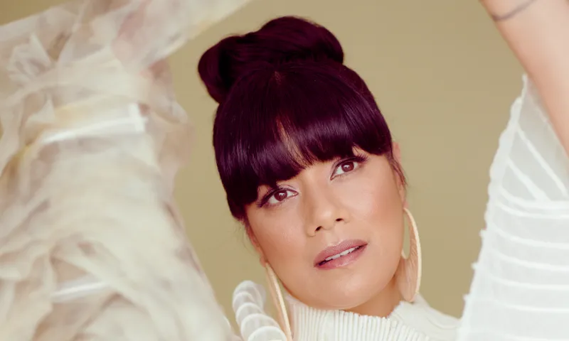 a close up portrait of a woman with black bangs and wearing her hair in a bun, large gold hoop earrings and a flowing cream dress