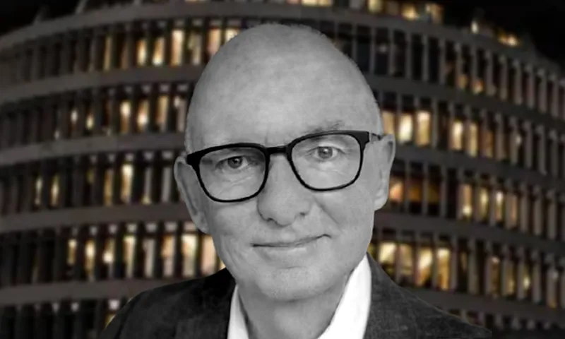 A man with glasses and a slight smile faces the camera, wearing a jacket and shirt. The background shows a multi-story building with many lit windows at night.