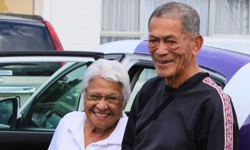 An elderly woman with white hair and glasses smiles beside an elderly man standing outdoors near parked cars, both appearing happy.