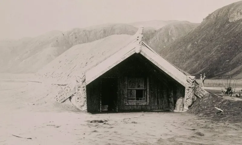 A Māori meeting house partially buried in volcanic ash, with intricate carvings on its facade, set against barren hills in the background.