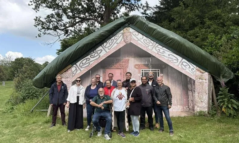 A group of people pose and smile in front of a traditional Māori meeting house set in a grassy outdoor area with trees and cloudy sky in the background.