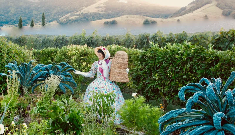 A woman in a floral dress and bonnet holds a beehive basket while tending to a lush garden with large leafy plants, green bushes, and hills in the background.