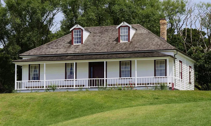 A small white house with a wooden shingle roof, dormer windows, a large front porch with railings, and a brick chimney, sits on a grassy hill with trees in the background.