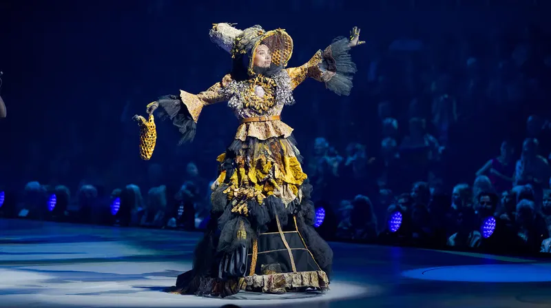 A performer on stage wears an elaborate costume with black and yellow layers, a large hat, and dramatic textures, surrounded by a seated audience in a dimly lit venue.