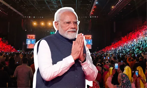 A man in a gray vest stands with folded hands in front of a large crowd at an indoor event with bright red and orange lights.