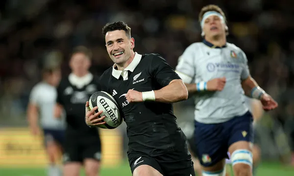 a rugby player in an all blacks uniform smiles while running with the ball as a player in a grey uniform chases behind