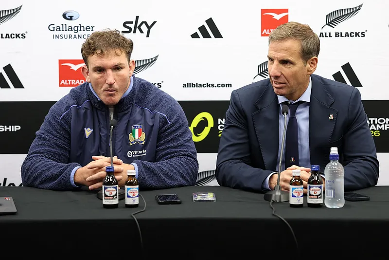 two men sit at a black table in front of a media screen and microphones, one wears a blue winter sports jacket and the other a blue suit and tie