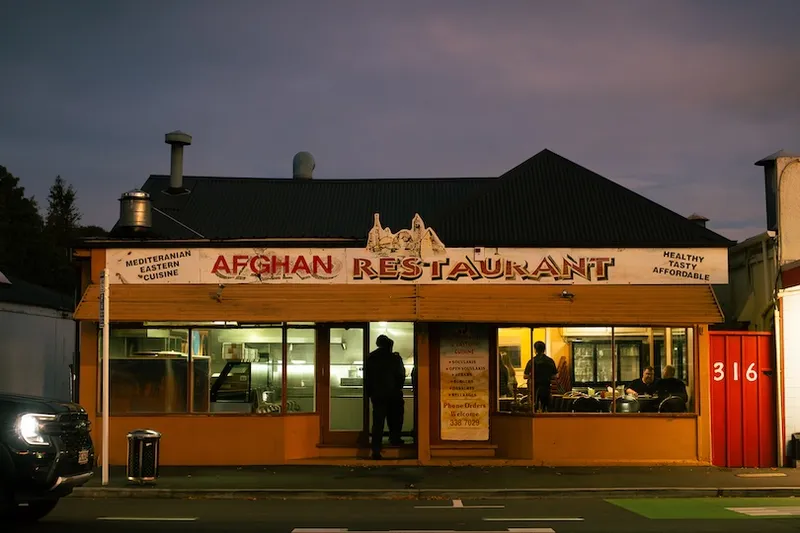 A warmly lit Afghan restaurant at dusk with large windows. One person stands outside, while others dine inside. The sign advertises Mediterranean and Eastern cuisine, and the words “Healthy, Tasty, Affordable” are visible.