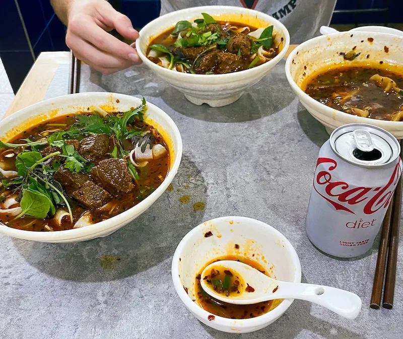 Three bowls of spicy beef noodle soup, a small empty bowl with a spoon, chopsticks, and a can of Diet Coke on a gray tabletop. A person’s hand is reaching for one of the bowls.