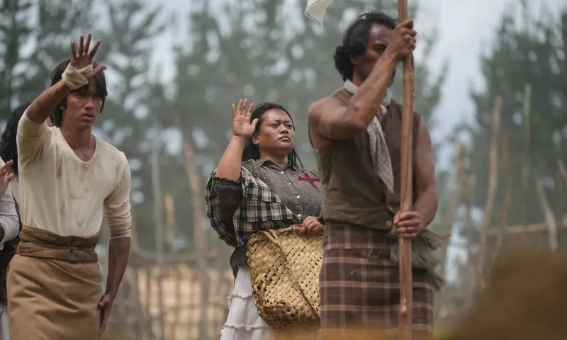 Three people in historical attire stand outdoors, raising their right hands. The woman in the center carries a woven basket. The background shows trees and wooden poles, suggesting a rural or historical setting.