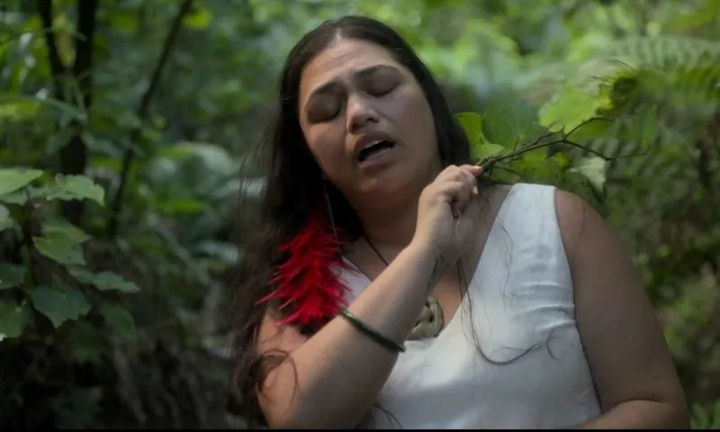 A woman in a white dress stands in a lush green forest, touching a leafy branch to her neck, eyes closed, with a red feather earring visible.