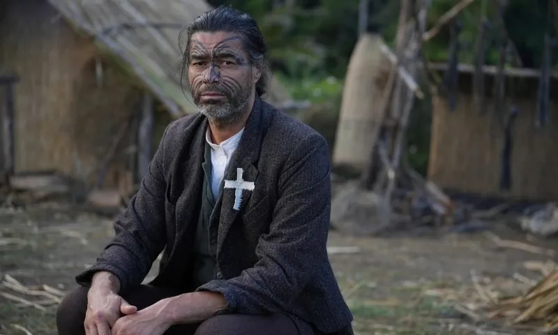 A man with dark facial markings, wearing a suit jacket and a white cross pinned to it, sits outside near traditional thatched huts and bamboo structures, looking seriously at the camera.
