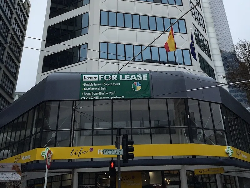 A commercial building displays a large green "For Lease" banner above glass windows. Two flags hang above, and the street sign reads "Victoria St." Yellow pharmacy signage is visible at street level.