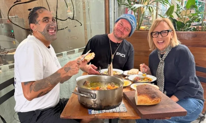 Three people sit at a table smiling, sharing a meal with bread and a large pot of food. The setting appears casual and cozy, with plants and wooden decor in the background.