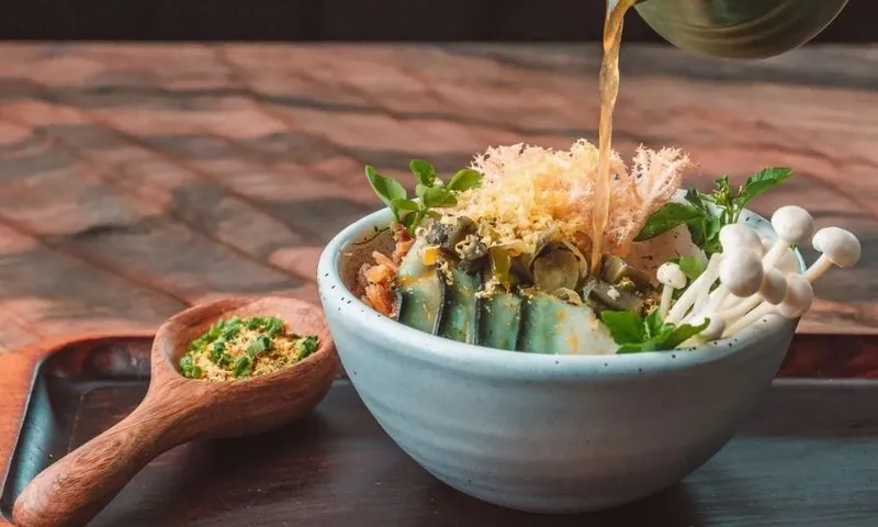 A bowl of boil up topped with vegetables, mushrooms, and herbs, with broth being poured in. A wooden spoon of seasoning sits beside the bowl on a wooden tray.