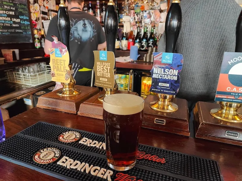 A pint of dark beer sits on a bar counter in front of three hand pumps labeled Reet Pale, Hillfoot Best Bitter, and Nelson Nectar. A bartender stands in the background near the taps.
