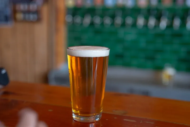 A pint glass filled with amber beer sits on a wooden bar with green tiles and beer taps blurred in the background.