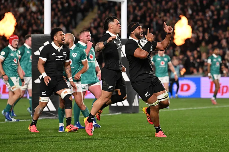 a group of rugby players wearing black uniforms celebrate together as players in green uniforms watch on and flames flare in the background