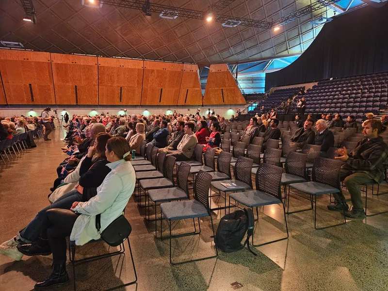 Audience members sit scattered in rows of chairs inside a large auditorium with high ceilings, empty seats, and warm lighting. The focus is on the sparse attendance and spacious setting.