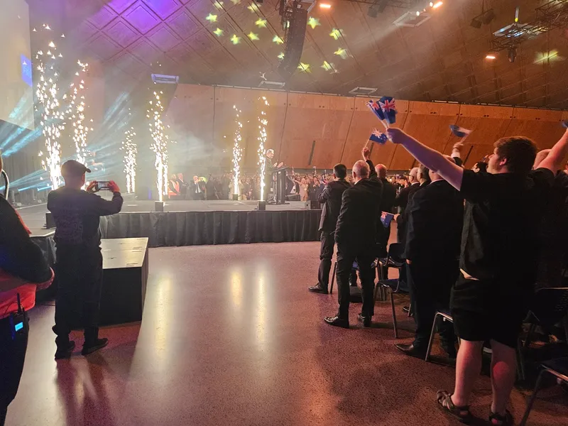 A crowd in formal attire cheers and waves flags at a stage event with bright lights and pyrotechnics, while a person on stage speaks at a podium.