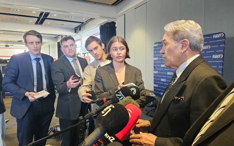A man in a suit speaks at microphones while four journalists, dressed formally, listen closely and take notes or record him in a conference room setting.