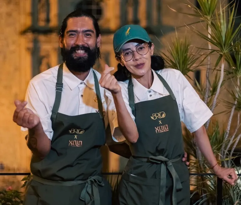 Two people wearing green aprons and white shirts stand together, smiling and making finger heart gestures. Plants are visible on the right, and the background is softly lit, suggesting an evening setting.