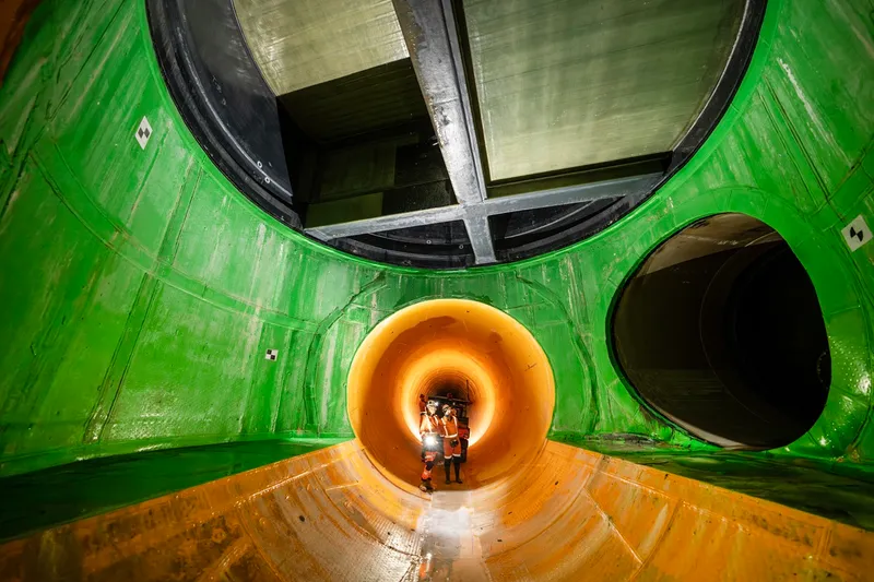 Workers in safety gear stand inside a large, brightly lit yellow-orange tunnel, with green and black circular structures surrounding the opening above them.