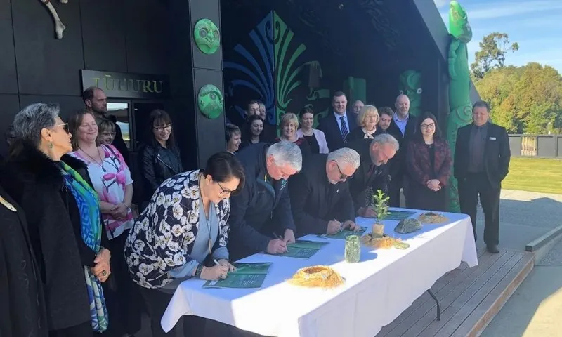 A group of people gathers outdoors while several individuals seated at a table sign documents. Māori carvings and decorations are visible on the building behind them. The mood appears formal and celebratory.
