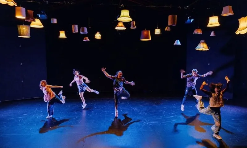 Five people dance energetically on a stage under many vintage lampshades hanging from the ceiling, with a dark background and dramatic lighting highlighting the performers.