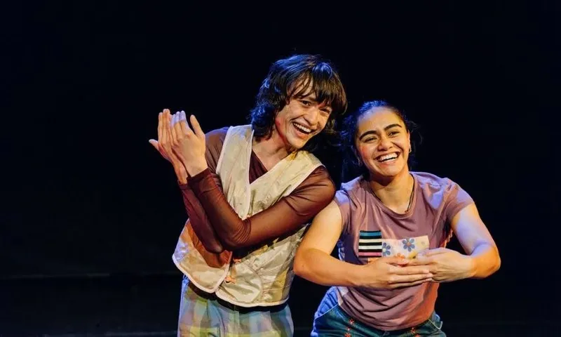 Two people stand together on stage, smiling and posing with their hands in front of them, as if miming an action. One wears a vest and plaid pants, the other a purple t-shirt with patches. The background is dark.