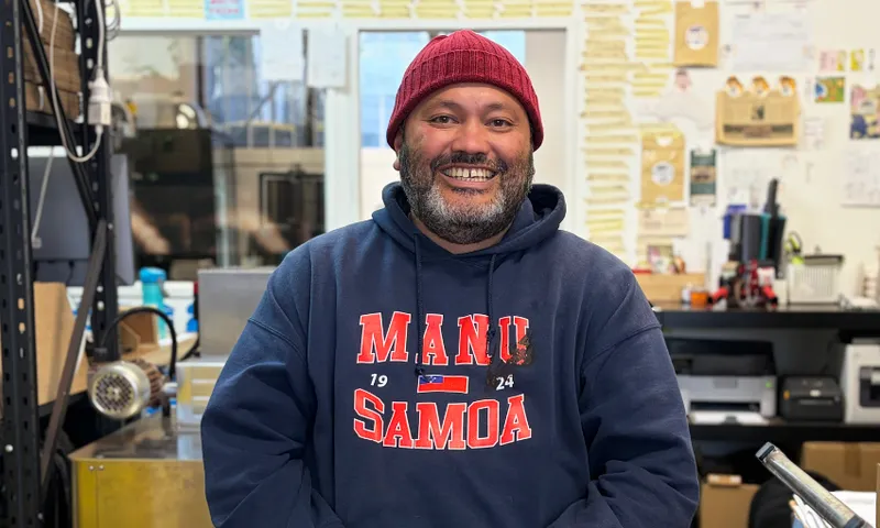 a man with brown skin and a black and white beard smiles at the camera wearing a red beanie and blue samoan hoodie. he sits in a cocoa bean roastery