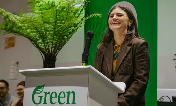 A smiling woman in a brown blazer stands at a podium labeled "Green," with a large fern and green backdrop behind her. A microphone is on the podium.