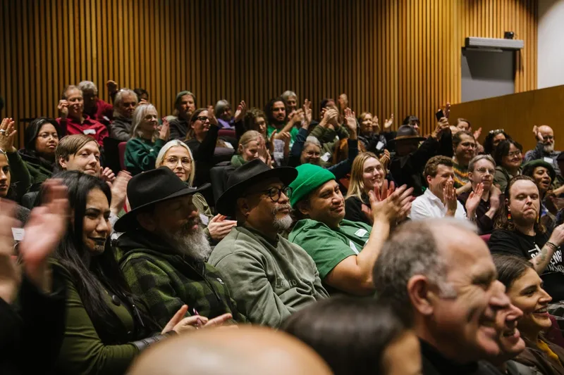 A diverse audience sits in rows, attentively watching an event. Many people are clapping, smiling, and dressed in casual attire. The background features vertical wooden paneling.