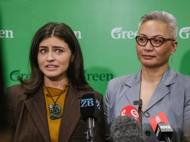 Two women stand in front of a green background with "Green" logos, speaking at microphones from news outlets. One woman looks concerned while the other looks calm and composed.