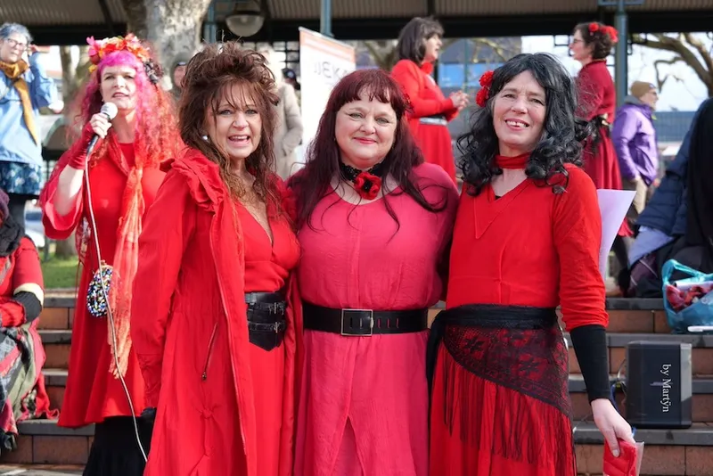Three women wearing red dresses and black belts are dressed as Kate Bush from Wuthering Heights. They stand closely together, smiling at the camera