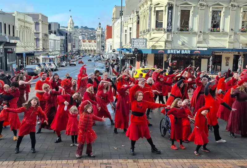 A large group of dancers wearing red, their arms lifted above their head. Behind them is the city of Dunedin
