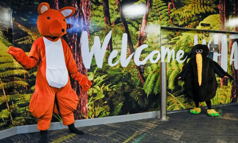 Two people in animal costumes—one as an orange kiwi and one as a black kiwi—stand in front of a wall mural with green ferns and the word “Welcome” on it.